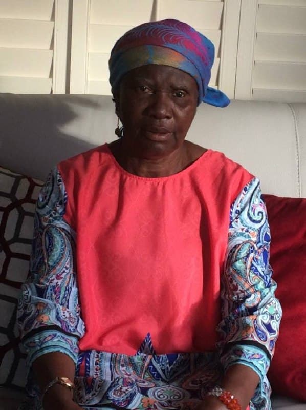 Mami in a soft pink blouse and headwrap, white shutters behind