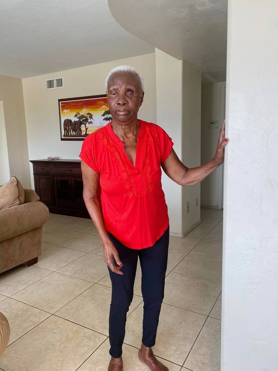 Mami in a red blouse, standing in the living room with an African sunset painting behind her